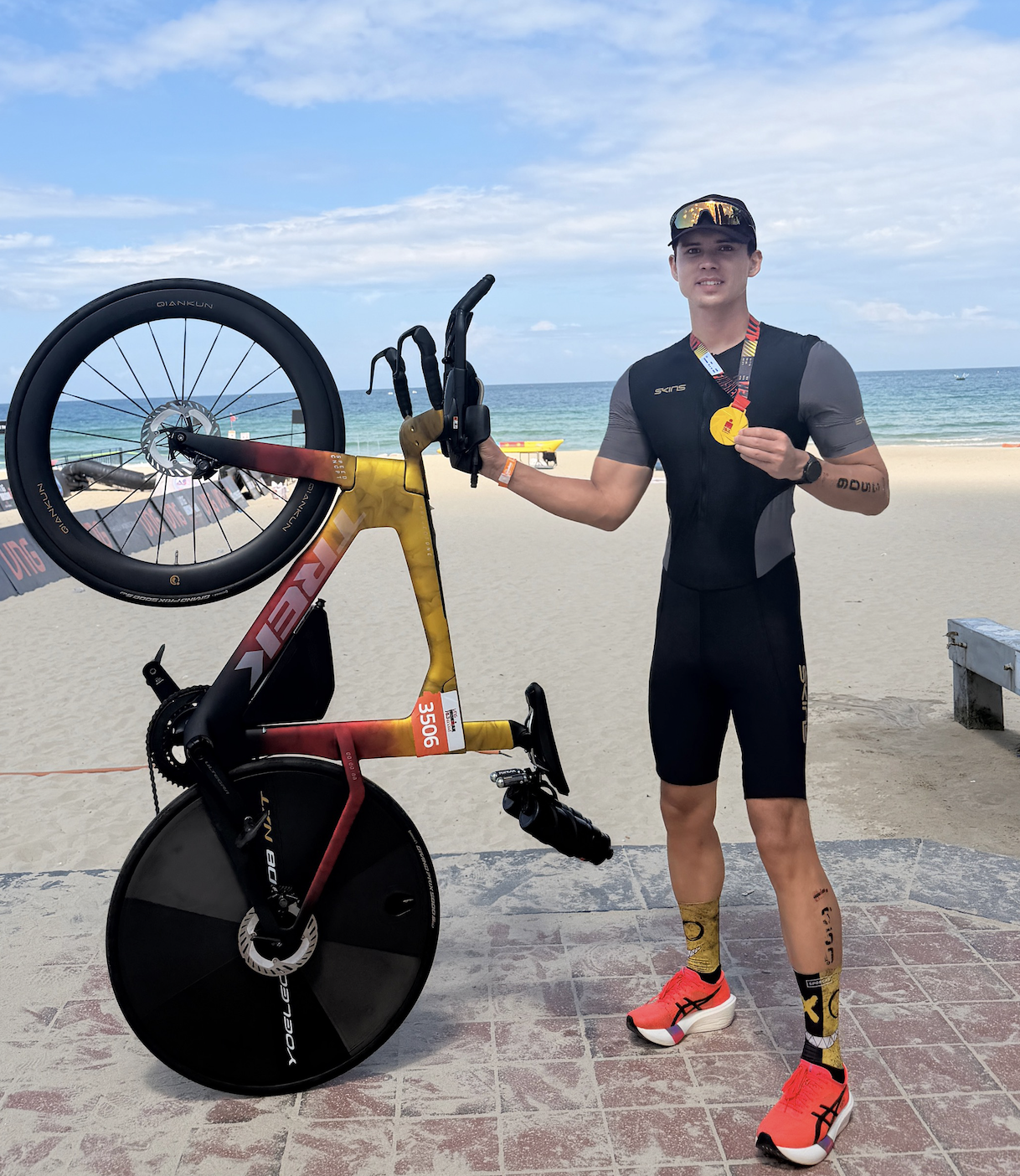 Mike Dee holding his triathlon bike and medal on the beach